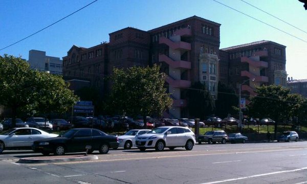 Large brick building with pink external stairs, bay windows, and trees, overlooking a street with cars.