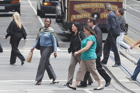 Pedestrians walk across a San Francisco street, with a cable car and vehicles in the background.