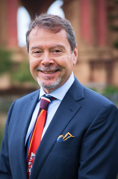 Claude W. smiles in a navy suit, patterned red tie, and pocket square, with a blurred arched building background.