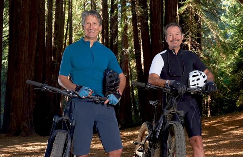 smiling people in cycling gear hold mountain bikes in a redwood forest.