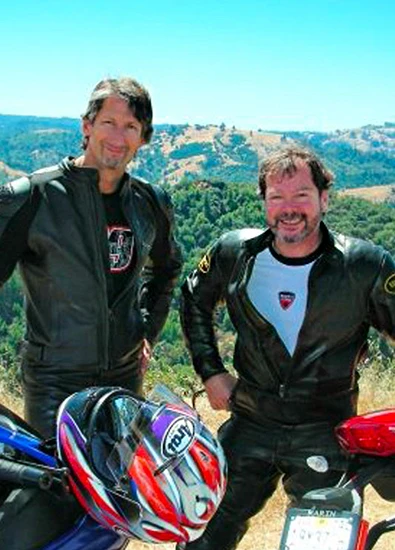 smiling people in motorcycle suits, one with a Ducati logo, stand with an Arai helmet and bikes, overlooking hills.