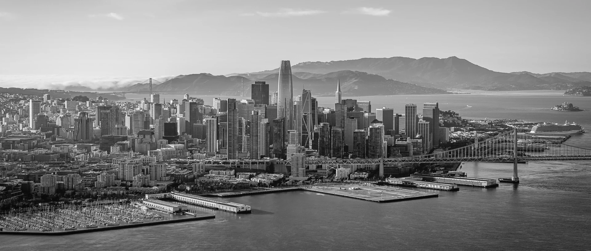 Black and white aerial view of San Francisco city skyline, waterfront, Bay Bridge, and distant mountains under a cloudy sky.
