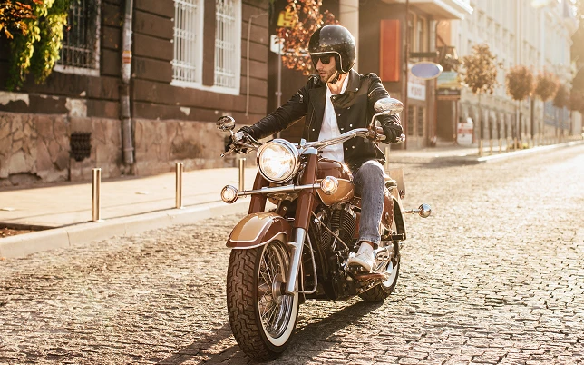 a vintage brown and gold motorcycle down a sunny cobblestone street, wearing a black helmet and leather jacket.