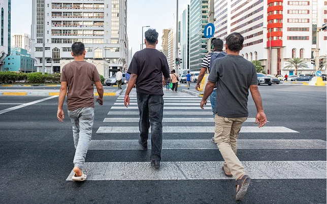 Four men walk away from the camera on a city zebra crossing, surrounded by tall buildings.