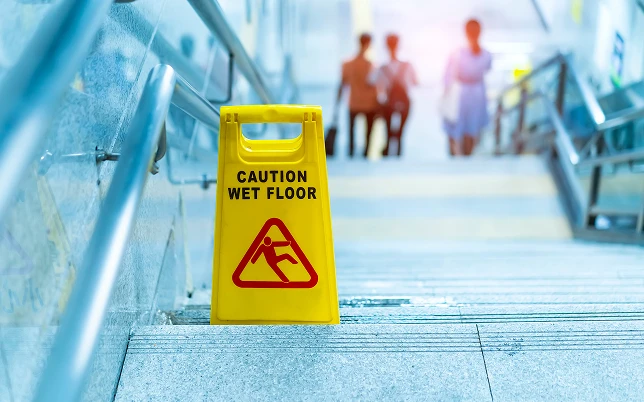 Yellow CAUTION WET FLOOR sign on grey stairs with blurry people walking and metal handrails in a blue-lit area.